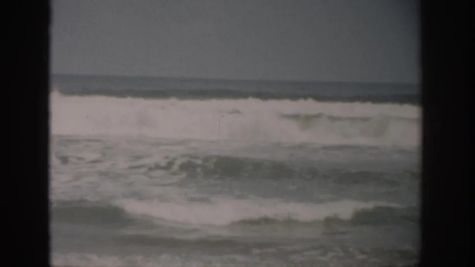 1959 Florida home-movie frame showing a low ocean wave breaking into white foam beneath a flat gray sky.