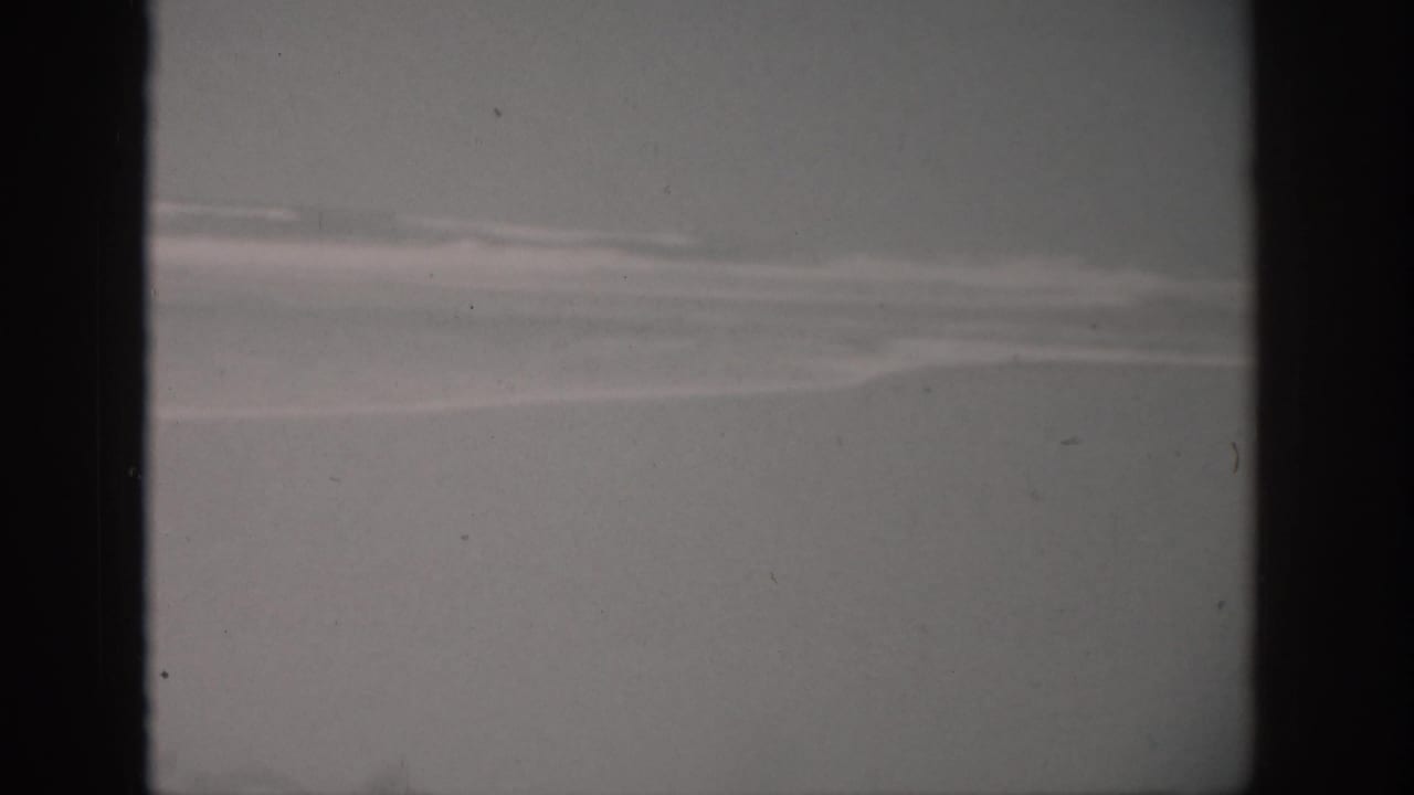 Distant surf lines over pale beach and water, framed by dark 8mm film borders.