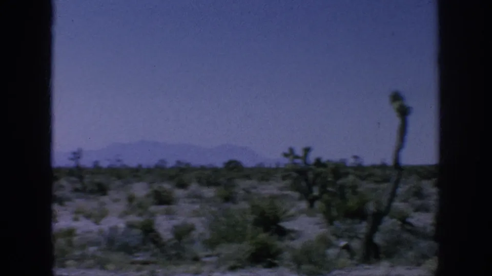 1962 8mm road-view frame showing arid scrubland, cactus-like plants, hazy sky, and distant low mountains through a window.