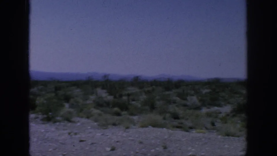 Soft 8mm frame of roadside arid terrain with low brush, pale ground, and a distant mountain ridge under a hazy sky.