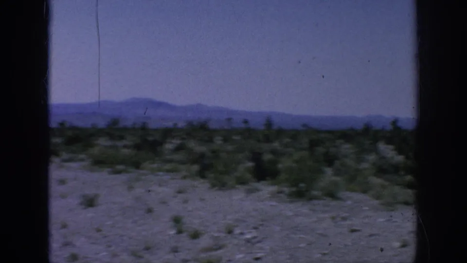 Arid landscape with scattered scrub, blue-toned mountain silhouettes, a dark window edge, and visible film scratches.