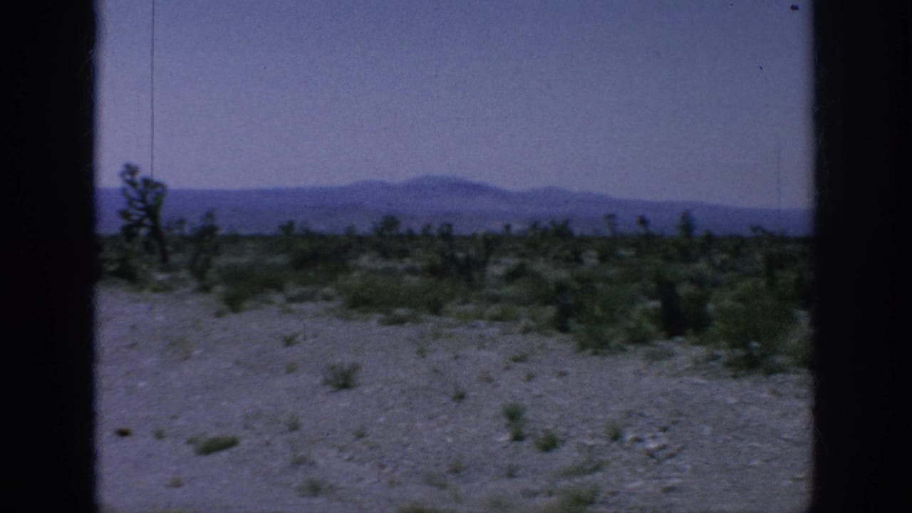 1960s 8mm road-view frame showing sparse roadside vegetation, pale sky, and distant mountains through visible gate borders.