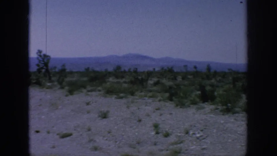 1960s 8mm road-view frame showing sparse roadside vegetation, pale sky, and distant mountains through visible gate borders.