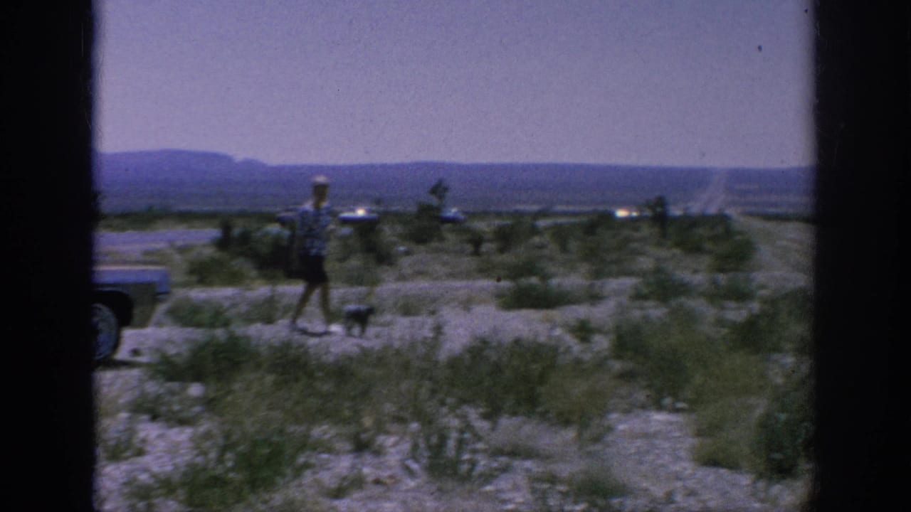 1962 8mm home-movie frame showing a traveler and small dog near a classic car with sparse vegetation and distant hills.