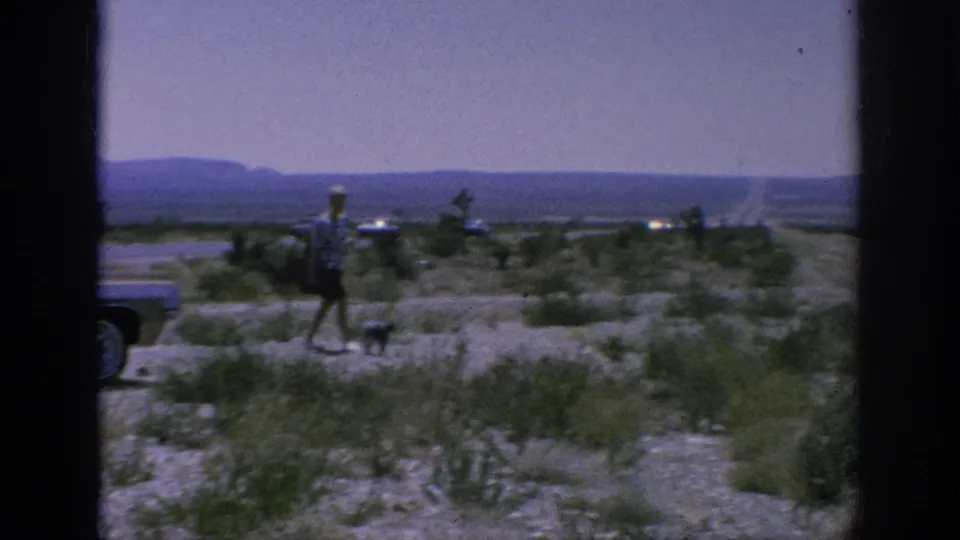 1962 8mm home-movie frame showing a traveler and small dog near a classic car with sparse vegetation and distant hills.
