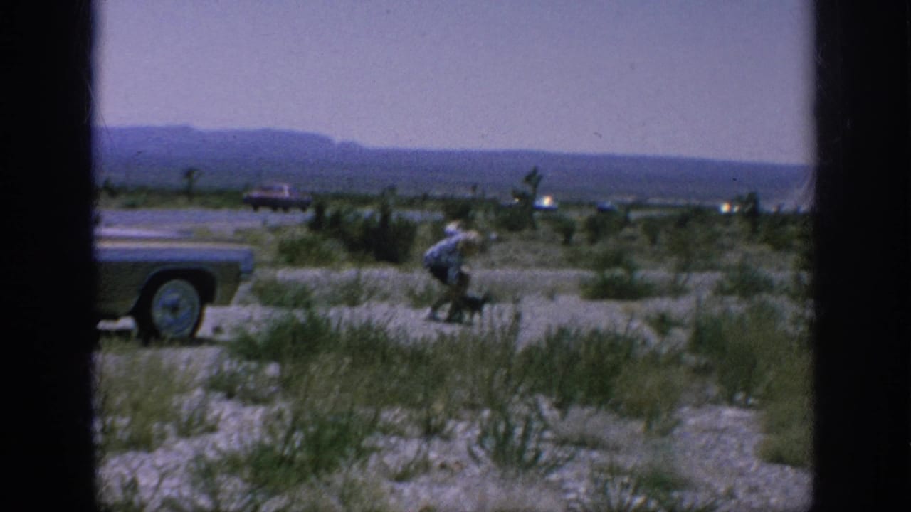 A 1962 home-movie frame shows a classic car at a roadside stop while a person bends near sparse vegetation.