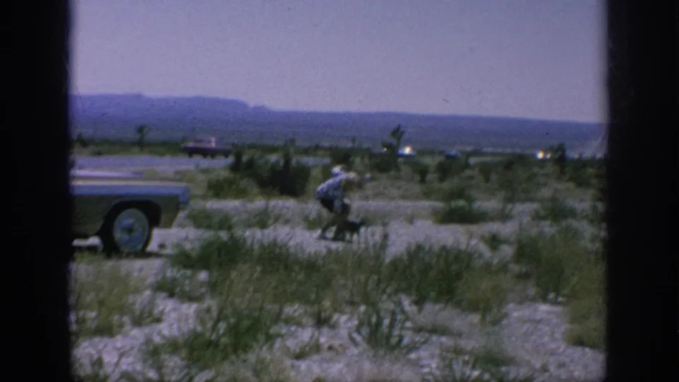 A 1962 home-movie frame shows a classic car at a roadside stop while a person bends near sparse vegetation.