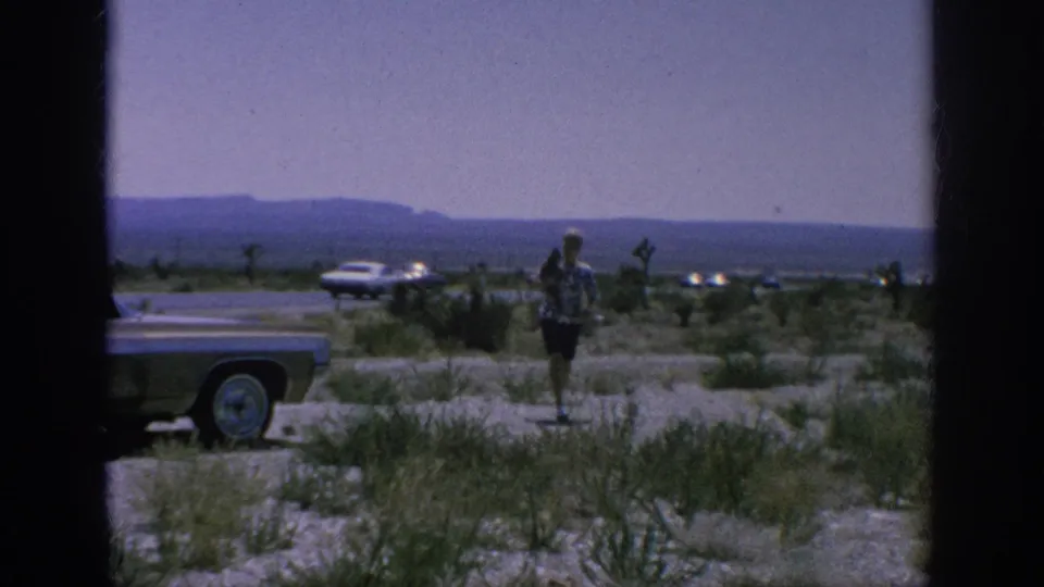 A traveler in a patterned shirt stands near a classic car in a dry open roadside setting, with distant vehicles and hills visible.
