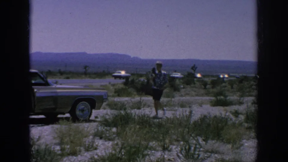A 1962 8mm frame shows a traveler beside a parked classic car, with sparse brush, distant road traffic, and hills beyond.