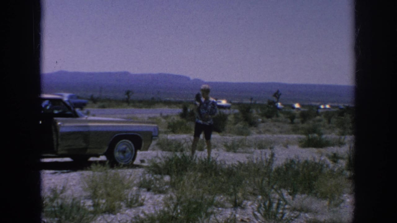 Woman standing beside a parked classic sedan on a gravel roadside with mountains in the distance.
