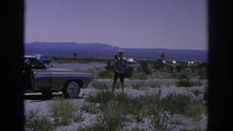 1962 8mm home-movie frame of a woman near a parked sedan on an open roadside, with passing cars, scrub vegetation, and distant mountains.