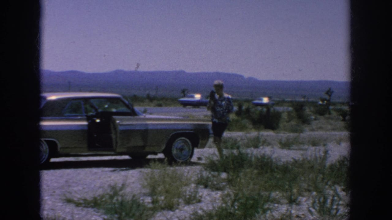 1962 8mm frame showing a woman beside a classic American sedan with the door open on a gravel shoulder under a hazy sky.
