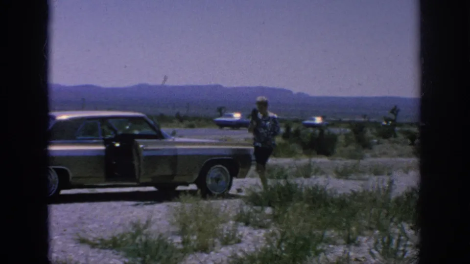 1962 8mm frame showing a woman beside a classic American sedan with the door open on a gravel shoulder under a hazy sky.