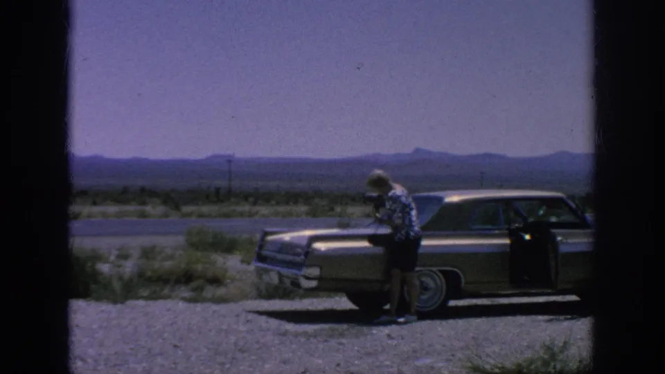 1962 home-movie frame of a woman bending near the rear of a parked sedan, with an open door, low vegetation, and distant mountains behind her.