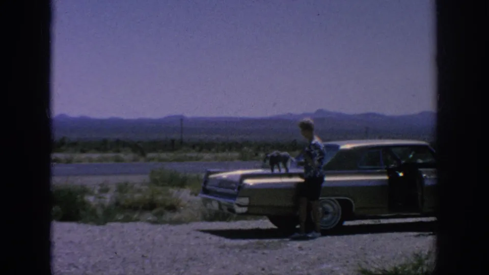 1962 8mm frame showing a woman beside a parked sedan with a cat visible on or near the car, set against a hazy roadside landscape.