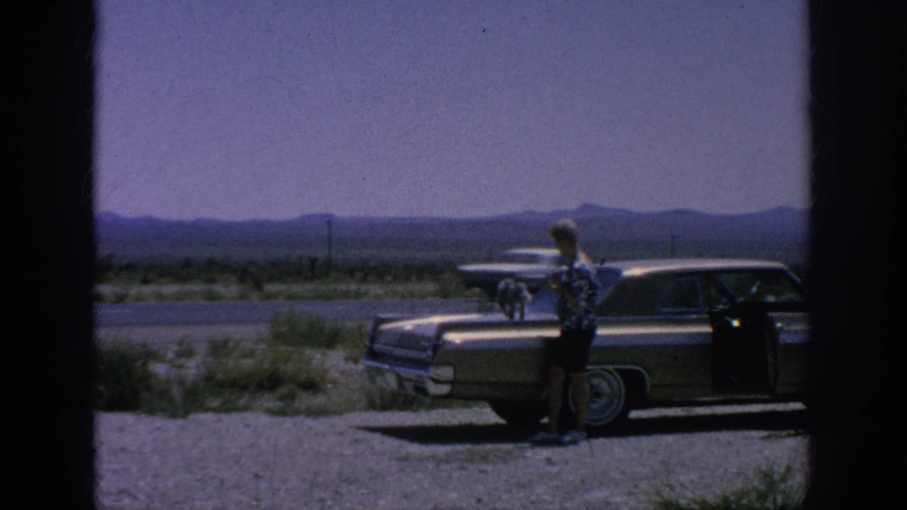 Man stands beside a parked sedan with a small dog on the hood near a desert highway.