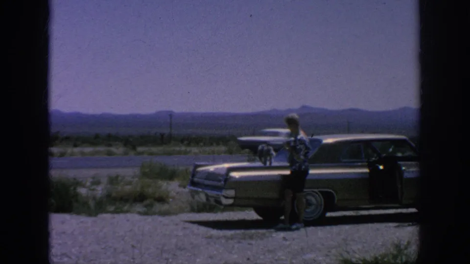 1962 home-movie frame of a man beside a classic sedan with a small dog on the hood along an open highway landscape.