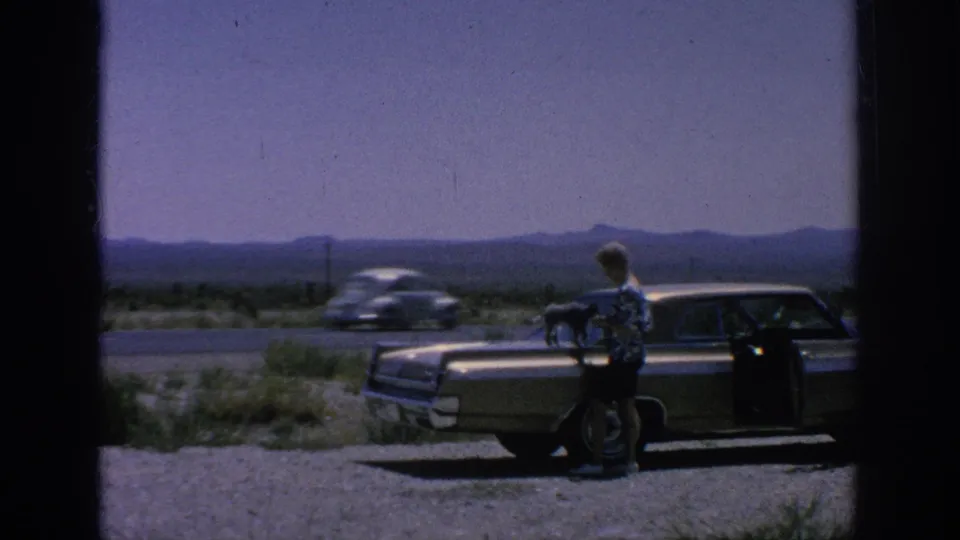 A white car passes behind the roadside sedan as the man stands near the dog on the hood in archival 8mm footage.