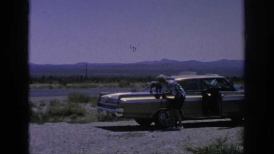 The man bends toward the small dog on the sedan hood during a brief 1962 home-movie roadside stop.