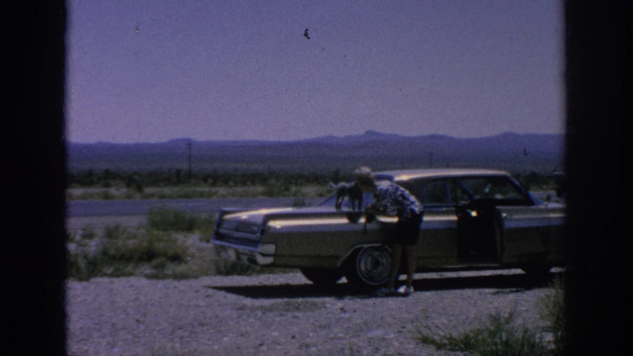 1962 home-movie frame showing a woman beside a classic sedan with a small dog on the hood along an open roadside.