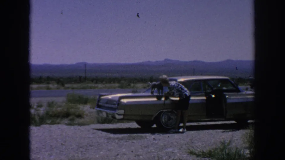 1962 home-movie frame showing a woman beside a classic sedan with a small dog on the hood along an open roadside.