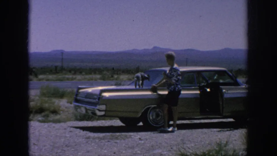 1962 home-movie frame showing a woman beside a classic sedan as a leashed poodle stands on the hood near a highway with mountains beyond.