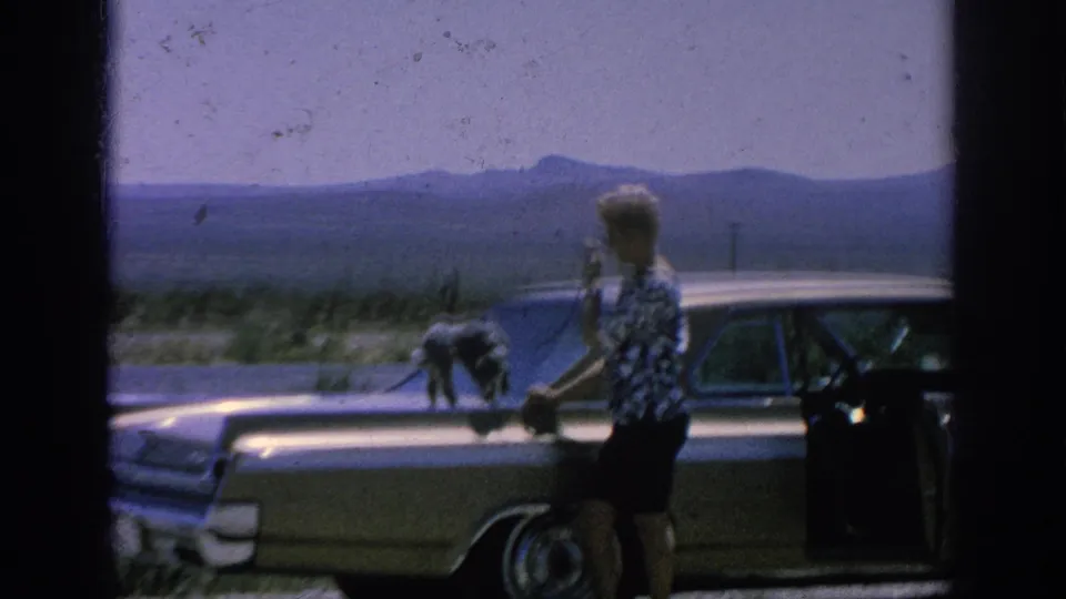 1962 8mm roadside view of a woman holding a poodle's leash beside a classic sedan, with desert scrub and mountains behind.