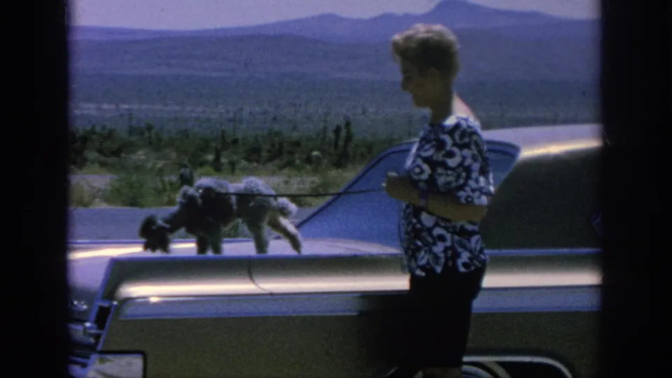1962 8mm frame of a woman beside a classic American car as a poodle stands on the trunk, with scrubland and distant mountains behind.