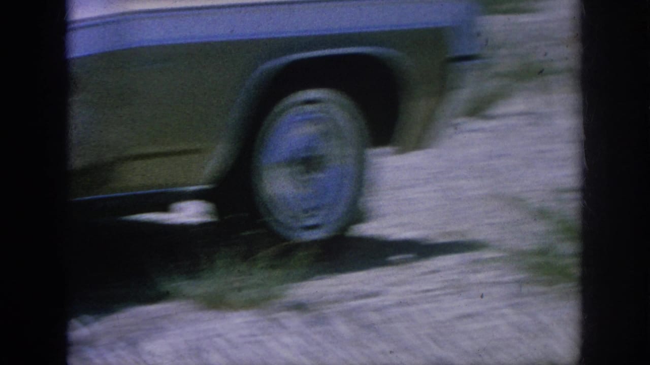 Soft 8mm frame showing the side and wheel of a classic car on a dirt shoulder, with blurred ground and vegetation at the edge.