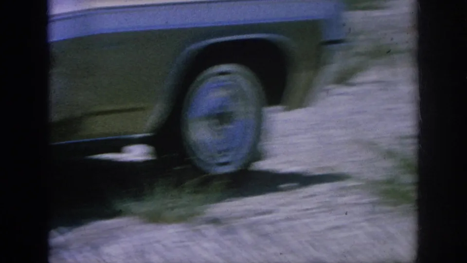 Soft 8mm frame showing the side and wheel of a classic car on a dirt shoulder, with blurred ground and vegetation at the edge.