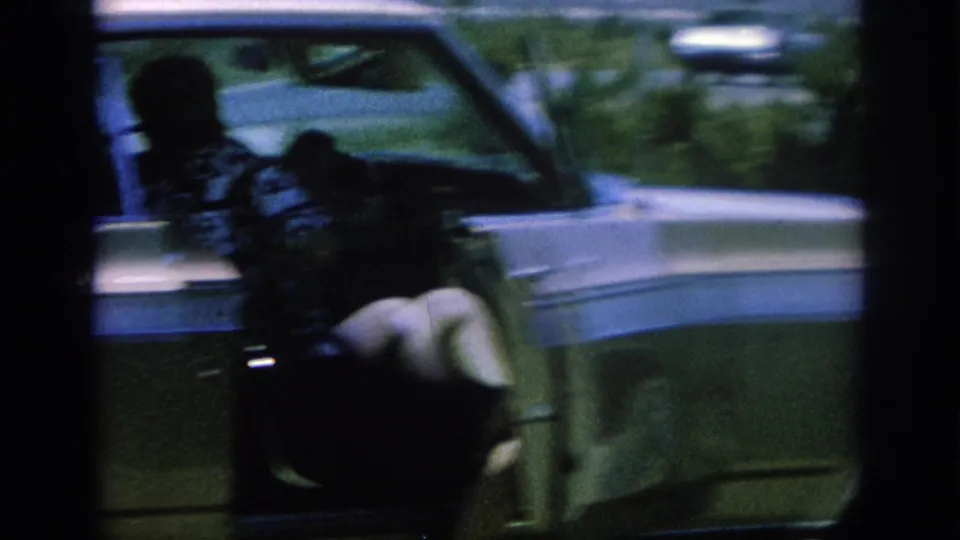 1962 8mm frame with heavy motion blur as a woman in a patterned blouse is seen at the side of a classic car near the open door area.