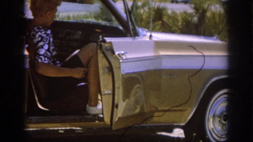 1962 8mm frame of a woman seated in a classic American car with the door open, parked near scrub vegetation in bright sun.