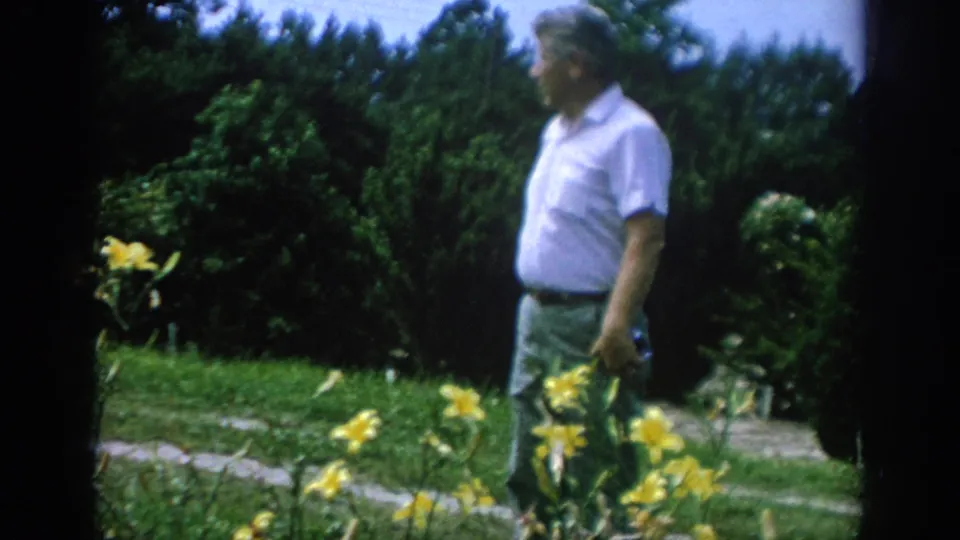 The man remains near the flower bed, with yellow blooms, lawn, shrubs, and trees visible in the 1962 Pennsylvania home-movie frame.