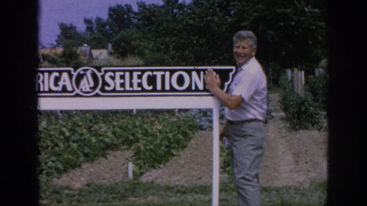 Smiling man with one hand on an All-America Selections sign by garden beds.