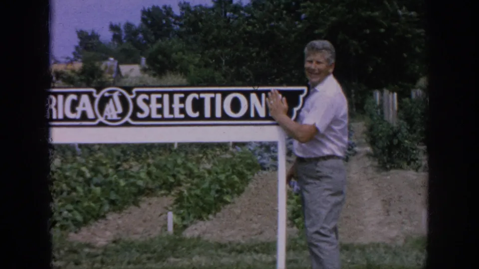 A smiling man poses with his hand on the All-America Selections sign in the 1962 Blainsport garden scene.