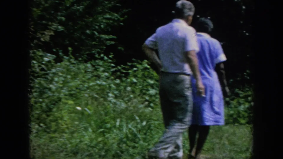 A man in a light shirt and a woman in a blue dress walk away through grass and dense greenery in the 1962 home-movie scene.