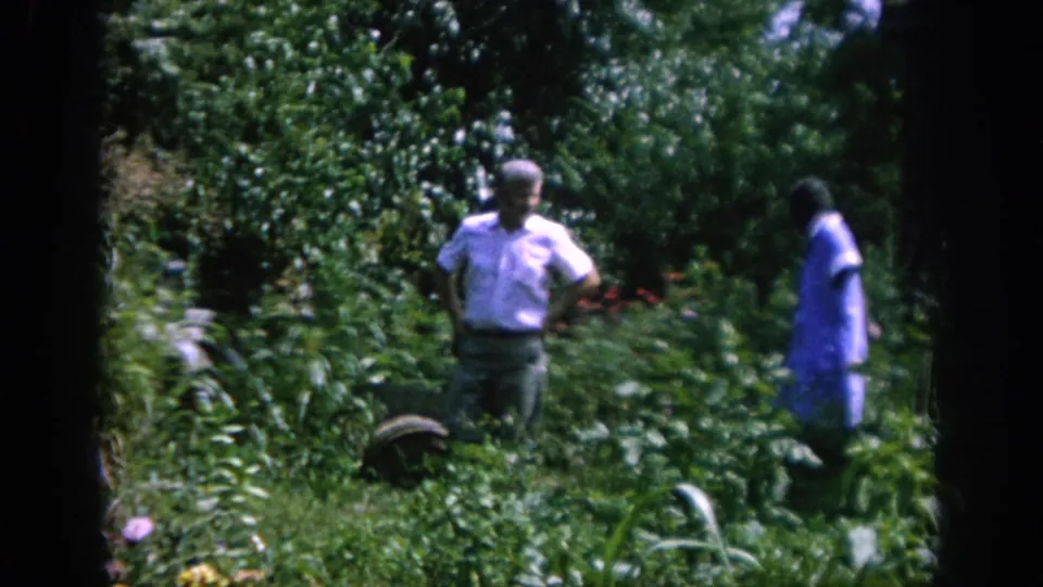 An older couple appears in a dense 1962 Blainsport garden, with the man in a light shirt and the woman in a blue dress.