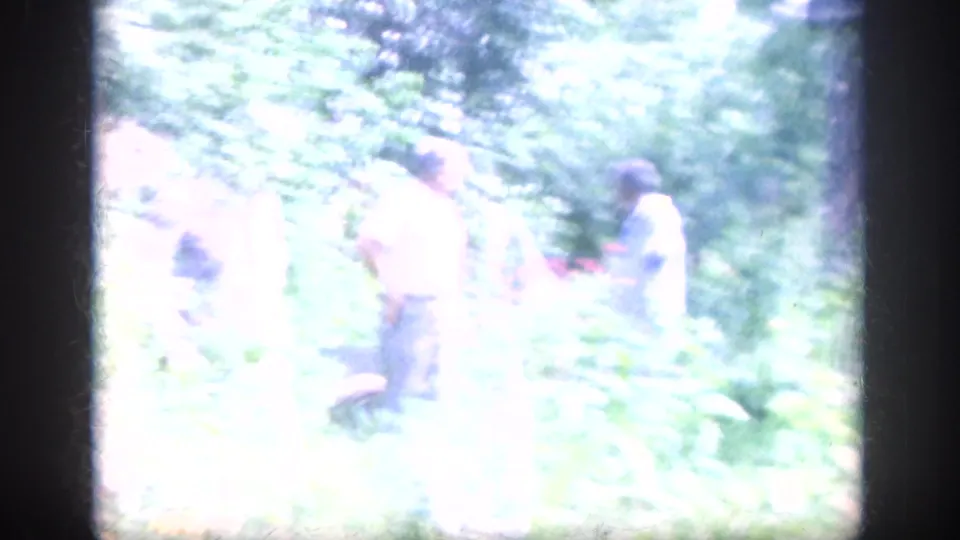 Bright, overexposed 8mm frame showing adults moving through the Blainsport garden amid pale sunlight and greenery.