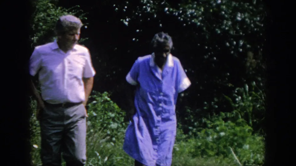 A man in a light shirt and a woman in a blue dress walk toward the camera through a leafy Blainsport garden scene.
