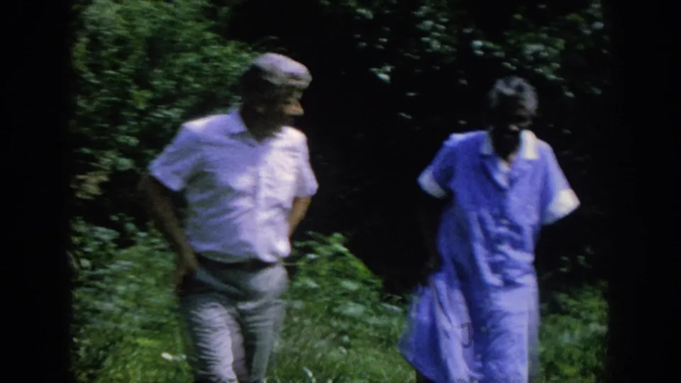 The two adults remain side by side as the handheld 8mm camera catches them moving through shaded garden foliage.