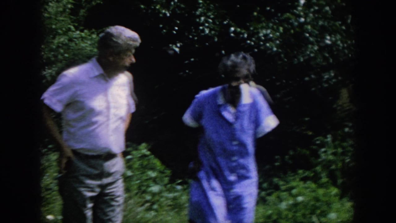 1962 Blainsport home-movie frame showing a man in a white short-sleeved shirt walking with an older woman in a blue dress outdoors.
