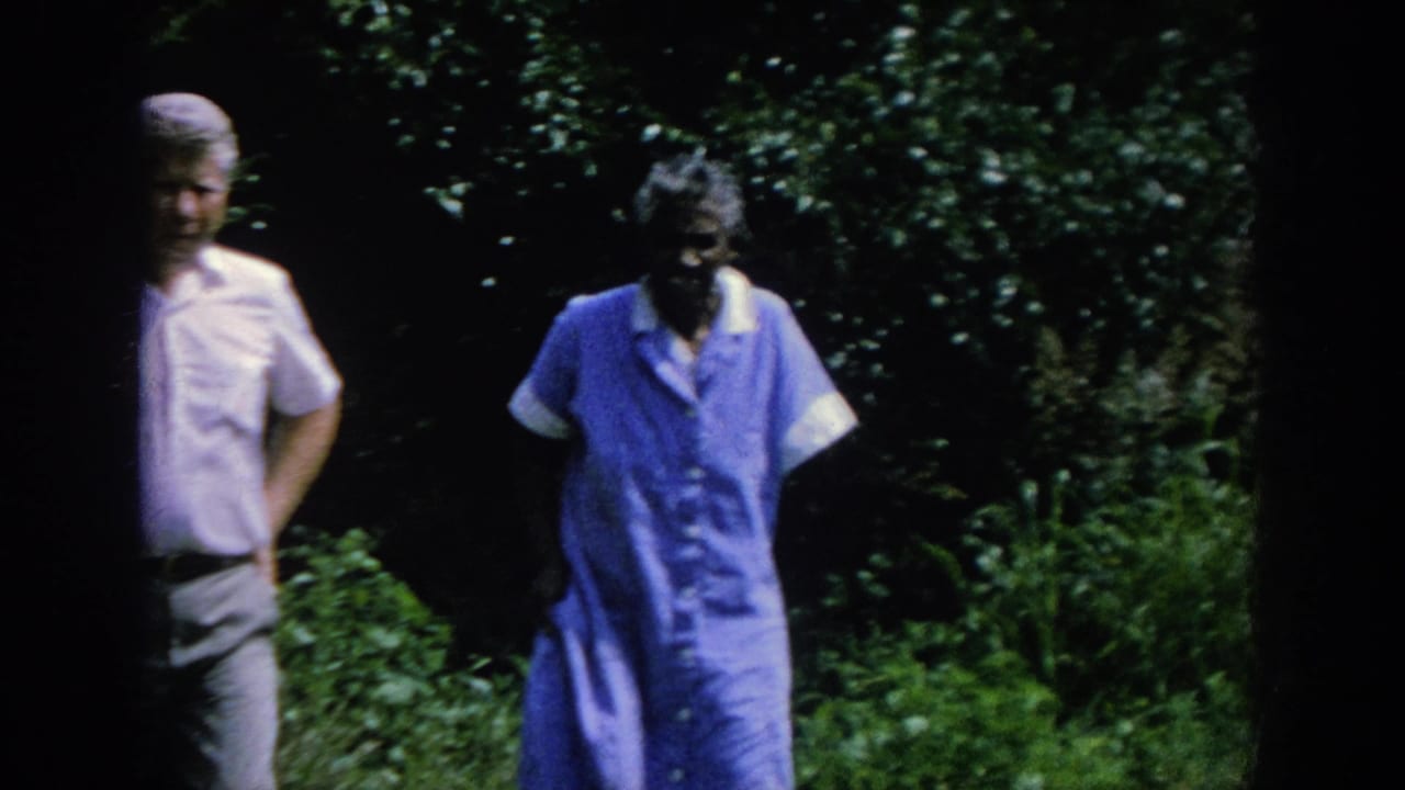 Man and older woman continue walking through a dark green garden area.
