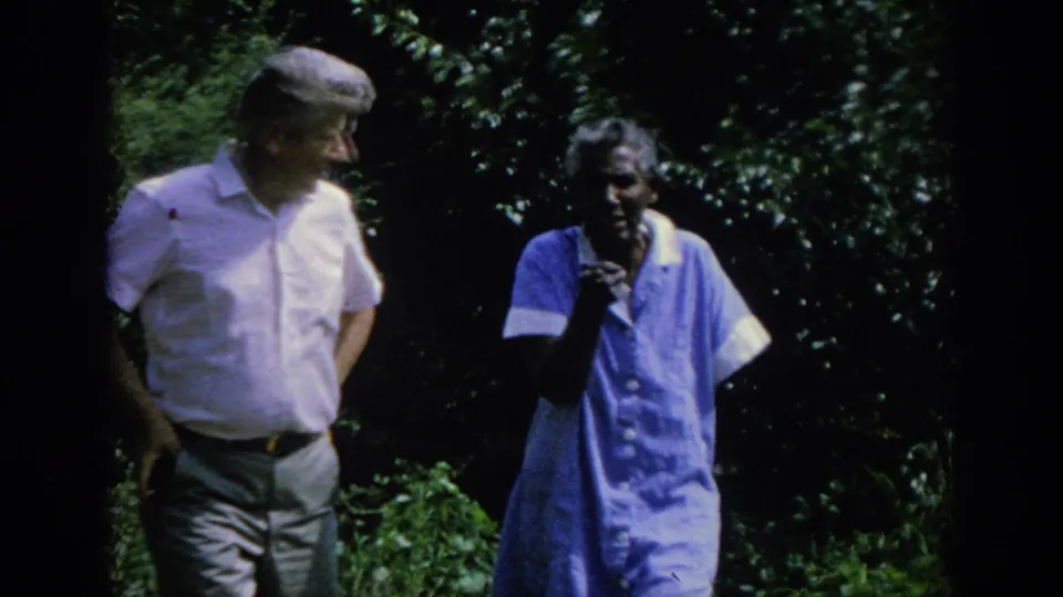 In this 1962 Blainsport frame, the older woman raises a hand while walking with the man through sunlit garden greenery.