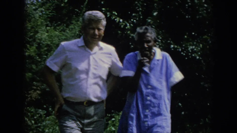 A 1962 Blainsport home-movie frame shows the man smiling as he walks with the older woman through a shaded green garden.