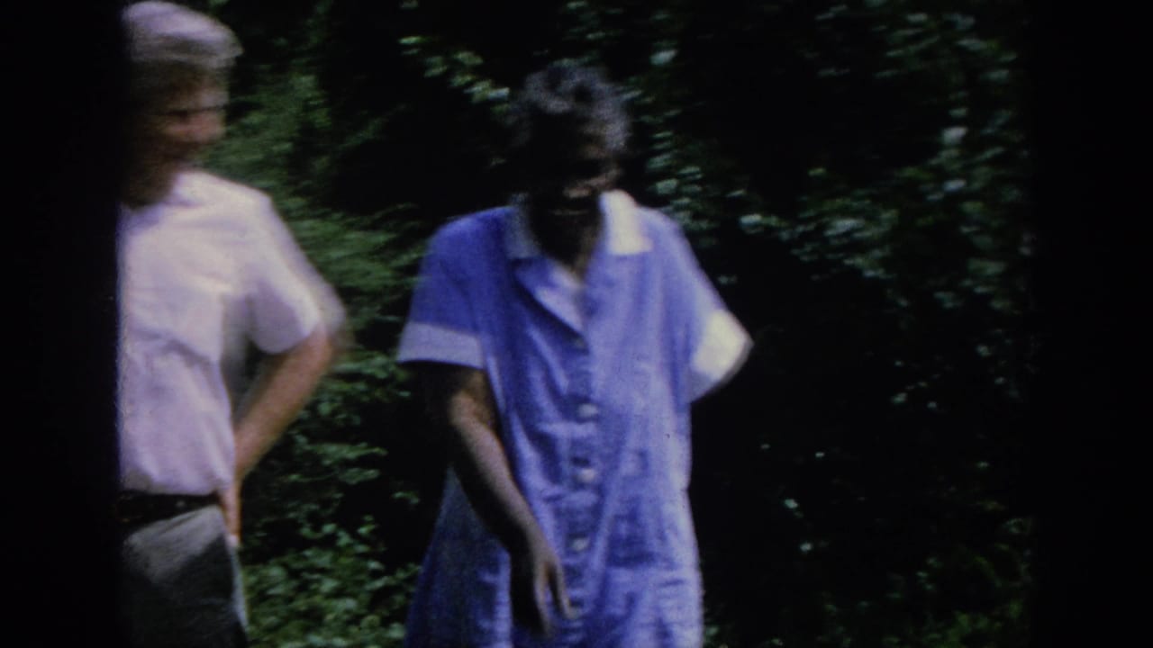 Man and elderly woman walk beside dense shrubs, partly blurred by motion.