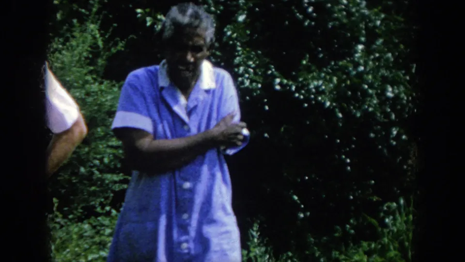 The elderly woman in a blue dress stands in front of dense greenery while the man is only partly visible at the edge.