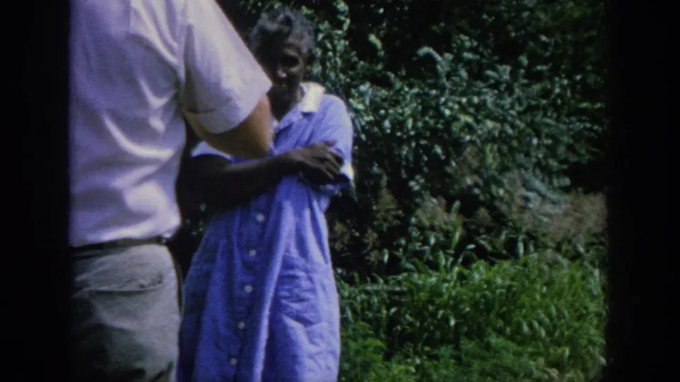 The man turns toward the elderly woman in the blue dress during an outdoor exchange in the Blainsport home movie.