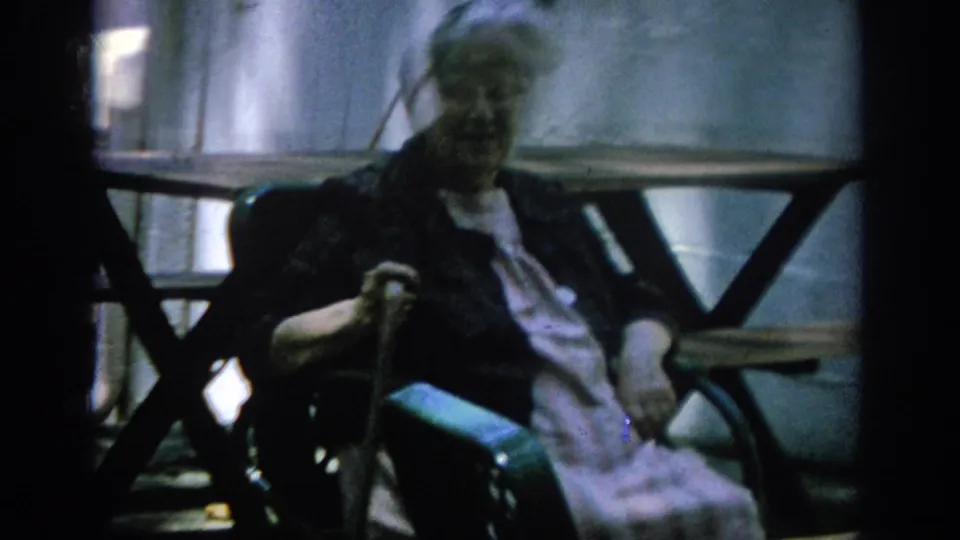 The elderly woman remains seated in her wheelchair in the Blainsport interior, with soft focus and a dark background around her.