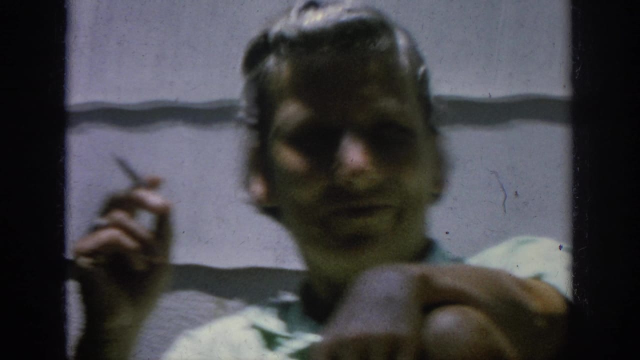 Man in a light shirt holds a cigarette near house siding and dark framing.