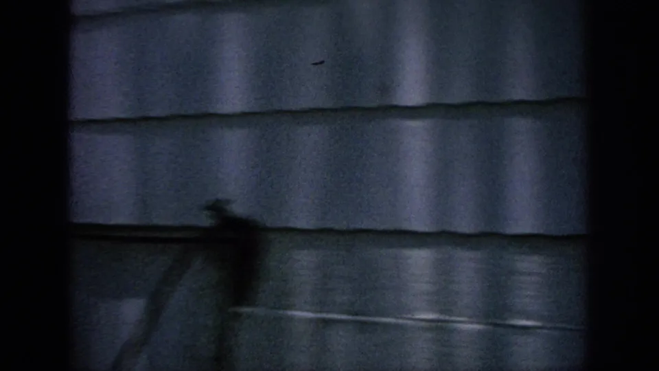 A dim 8mm frame shows exterior house siding through dark framing, with a blurred object or shadow near the wall.